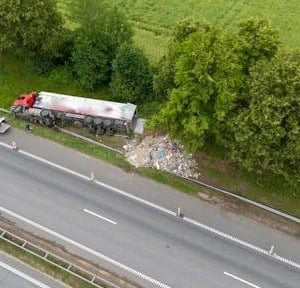 Aerial view of a red semi-truck that has left the road and come to a stop beside trees, with debris scattered on the grass.