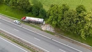 Aerial view of a road accident with a red truck overturned on the side, debris scattered, and a car parked nearby on a highway surrounded by trees and grass.