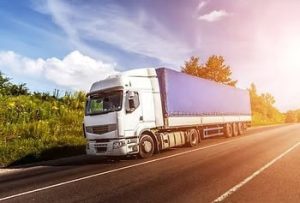 A white and blue cargo truck drives on a sunny rural road, surrounded by trees and grass, with a clear blue sky overhead.
