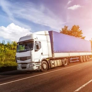 A white semi-truck with a blue trailer is parked on the side of a road on a sunny day, with green vegetation and trees in the background.