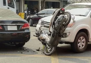 A motorcycle is wedged upright between a black car and a white SUV after a traffic accident on a city street.