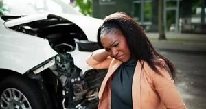 A woman stands with a pained expression, touching her neck, in front of a damaged white car on a street.