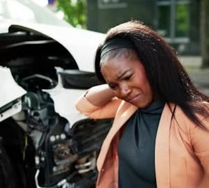 Woman standing beside a damaged car, holding her neck and appearing to be in pain or discomfort after an accident.