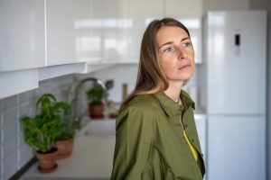 Woman with long hair in a green jacket stands pensively in a kitchen with white cabinets and a visible refrigerator and plants.