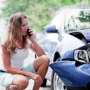 A woman crouches next to a damaged blue car, talking on her phone, appearing to report or seek help after a car accident.