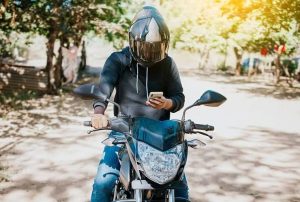 Person on a motorcycle wearing a helmet, looking at a smartphone, in an outdoor setting.