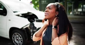 A woman, standing in front of a damaged white car, touches her neck, appearing to be in discomfort.