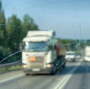 Photo taken through a vehicle windshield with visible cracks and chips; blurred trucks and road traffic appear in the background.