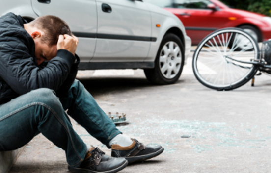 A man sits on the ground holding his head near a car and a fallen bicycle, with broken glass scattered on the pavement.
