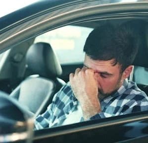 A man in a plaid shirt sits in the driver's seat of a car, resting his head on his hand and appearing stressed or tired.