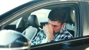 A man in a plaid shirt sits in a car, rubbing his eyes with his hand, appearing tired or stressed.