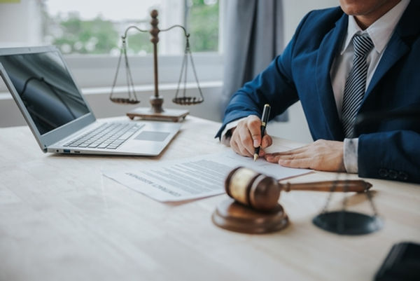 A person in a suit signs documents at a desk with a laptop, gavel, and scales of justice.