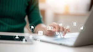Person using a laptop, touching a holographic icon labeled "TAX" among other finance-related symbols. Tablet visible in the foreground.