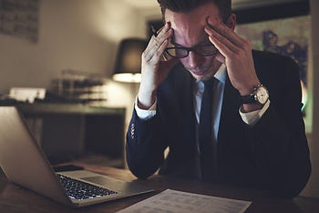 A man in a suit sits at a desk with a laptop, holding his head in his hands and looking stressed while reading documents.