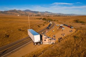 A semi-truck is stuck off a rural road with people gathered nearby. Several cars are parked along the road, and the landscape features dry grass and distant hills under a clear blue sky.