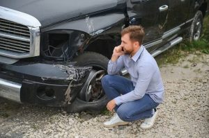 A man kneels next to a damaged black truck, examining the dented front left side and wheel.