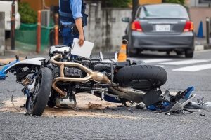 A motorcycle lies on its side on a road with scattered debris around it, while a person in a blue uniform stands nearby holding a document. A car is parked in the background.