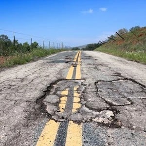 A rural road with a double yellow line is heavily damaged by potholes and cracks, with grass and a fence on either side under a clear blue sky.