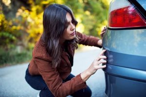 Woman in a brown jacket examines a scratch on the rear bumper of a blue car on a sunny day.