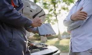 Two people stand in front of a car with an open hood. One holds a tablet, apparently discussing car issues. Trees and sunlight are visible in the background.