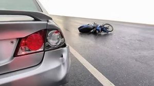 A silver car with a dented rear bumper is parked near a fallen blue motorcycle on a road.