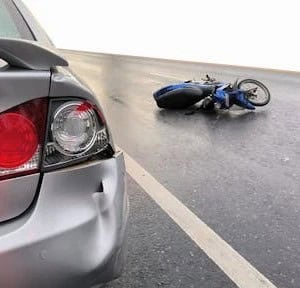 A damaged car and a fallen motorcycle are on a wet road, indicating a recent collision between the two vehicles.