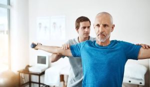 A man in a blue shirt is assisted by another man during a physical therapy session, stretching his arms out horizontally in a bright room.