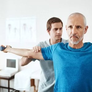 A physical therapist assists an older man in a blue shirt with a shoulder exercise in a clinical setting.