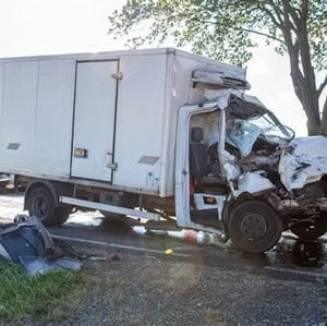 A white box truck with severe front-end damage is stopped on a road after a collision, with debris and fluid visible on the ground nearby.