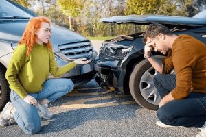 Two people kneeling near two cars with front-end damage after a collision. The woman gestures while the man holds his head.