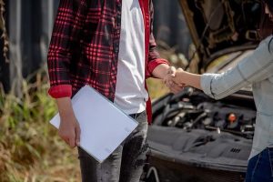Two people shaking hands in front of a car with an open hood. One person holds a clipboard.