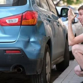 A man crouches beside a parked blue car with a noticeable dent on its rear left side, holding his head in apparent frustration.