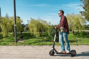 A person wearing a red plaid shirt and jeans rides an electric scooter on a paved path with green trees and grass in the background.
