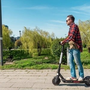 Man wearing sunglasses and a plaid shirt rides an electric scooter on a paved path in a park with green trees and grass under a clear blue sky.