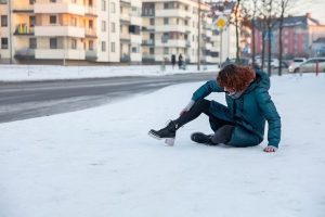 Person sitting on a snowy sidewalk after slipping, wearing a blue coat and black boots, with buildings and a road in the background.