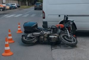 Motorcycle lies on its side next to a white van, surrounded by traffic cones, on a street.