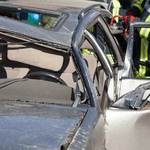 A silver car with a crushed roof and door sits damaged, while emergency responders in reflective gear work nearby.