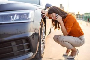 A woman in an orange blouse and beige pants squats near a car, holding a smartphone to her ear, inspecting the front wheel. The setting appears to be an outdoor parking area.
