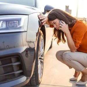 Woman crouching by a car with a flat tire, inspecting the damage while talking on a mobile phone.