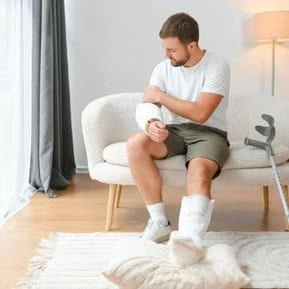 A man with a bandaged arm and leg sits on a sofa beside crutches, looking at his injured arm.