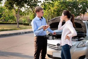 A man holding a clipboard talks to a woman in front of a car with an open hood on a sunny day. Trees and a street are in the background.