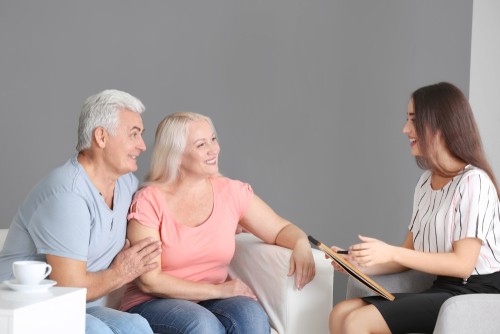 Two older adults sit on a couch and talk with a young woman who is holding a clipboard, in a casual indoor setting.