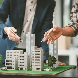 Two people point pens at a scale model of modern buildings, discussing or planning an urban development project.
