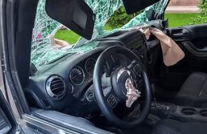 Interior view of a car with a shattered windshield and deployed airbags, showing significant damage to the dashboard area.