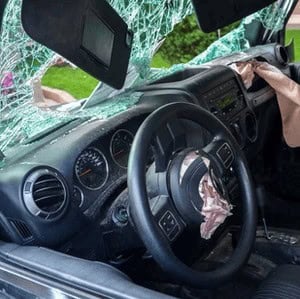 Car interior with shattered windshield, deployed airbags, and visible damage to the dashboard and steering wheel.