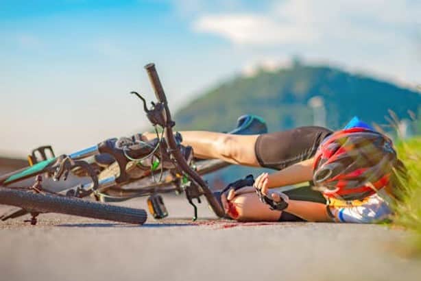 A cyclist wearing a helmet lies on the ground next to a fallen bicycle on a paved road, with a grassy hill in the background.