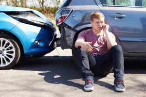 A man sits on the ground beside two cars involved in a rear-end collision, talking on a mobile phone.
