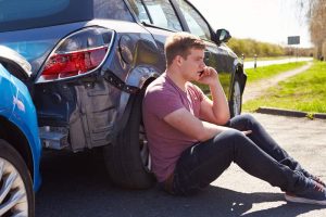 A man in a red shirt sits on the ground next to a damaged car, talking on a phone, with another vehicle nearby on a sunny road.