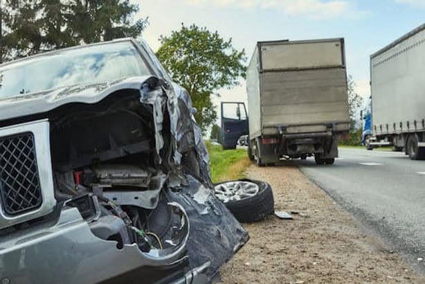 Front of a damaged SUV beside a road, with a detached wheel nearby and two trucks parked on the roadside in the background.