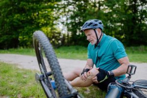 Older man in cycling gear sits on grass next to bike, holding his knee with a pained expression.